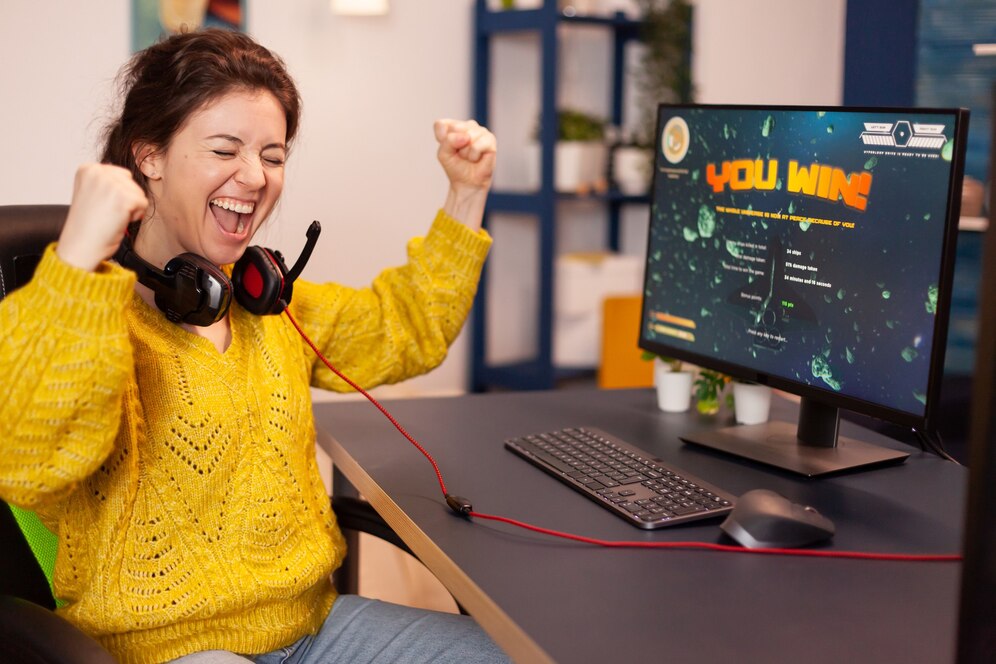 A person in a yellow sweater celebrates a gaming victory with raised arms, seated at a desk with a computer displaying YOU WIN.