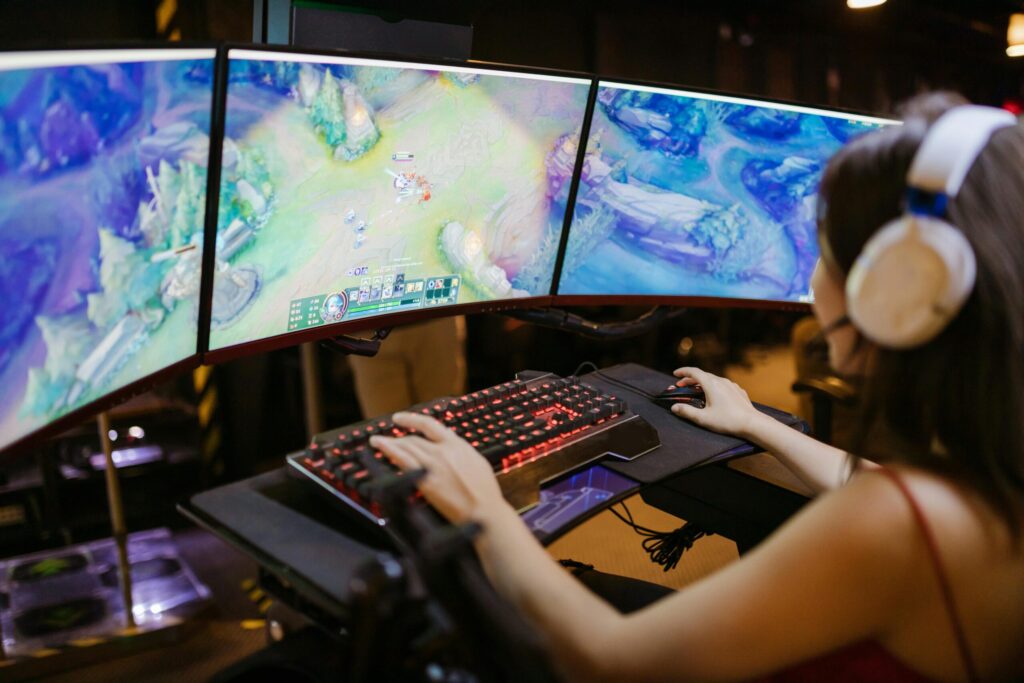 A gamer intently plays on a triple monitor setup, with a mechanical keyboard and headphones, immersed in a colorful game environment.