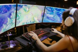 A gamer intently plays on a triple monitor setup, with a mechanical keyboard and headphones, immersed in a colorful game environment.