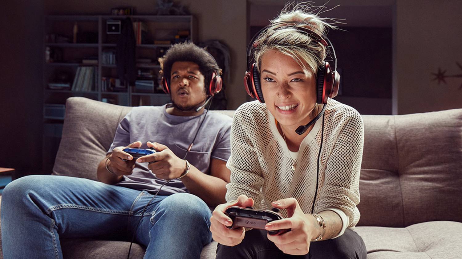  Two individuals wearing headsets are sitting on a couch, focused on playing video games with controllers in their hands.