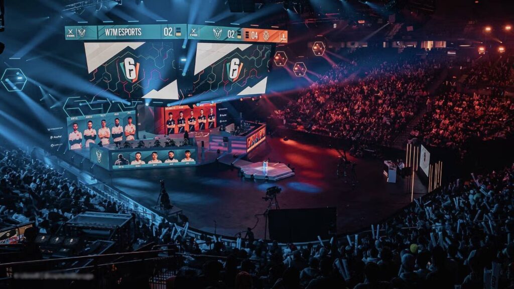 A vibrant esports arena packed with fans, featuring teams competing on stage, highlighted by a large digital scoreboard.