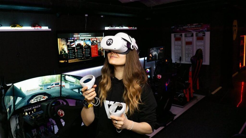 A person holds VR controllers in a gaming setup with racing screens and a racing simulator in a dimly lit room.