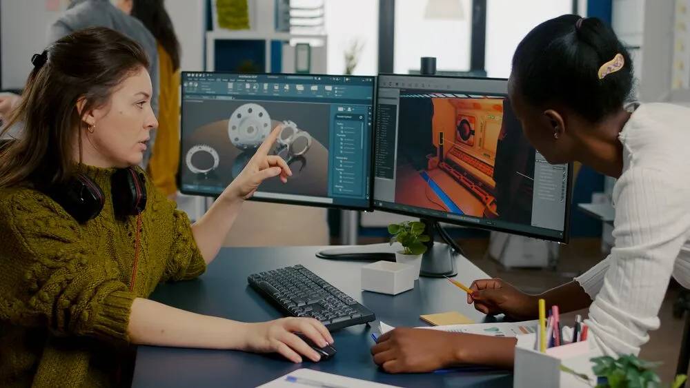 Two women work collaboratively at a desk with dual monitors displaying 3D modeling and design software, surrounded by office supplies.