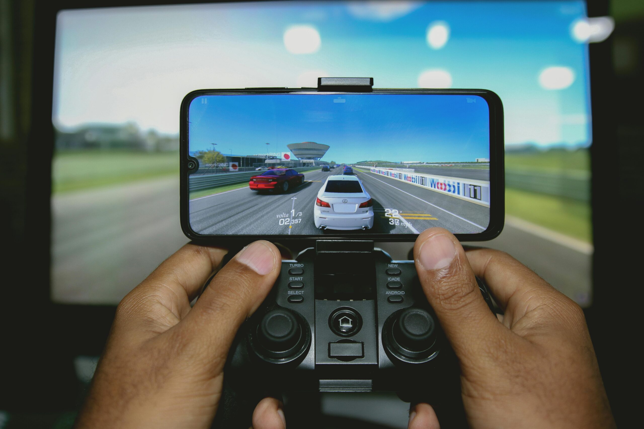 A gamer holds a mobile phone with a racing game displayed, while a larger screen shows the same gameplay in the background.