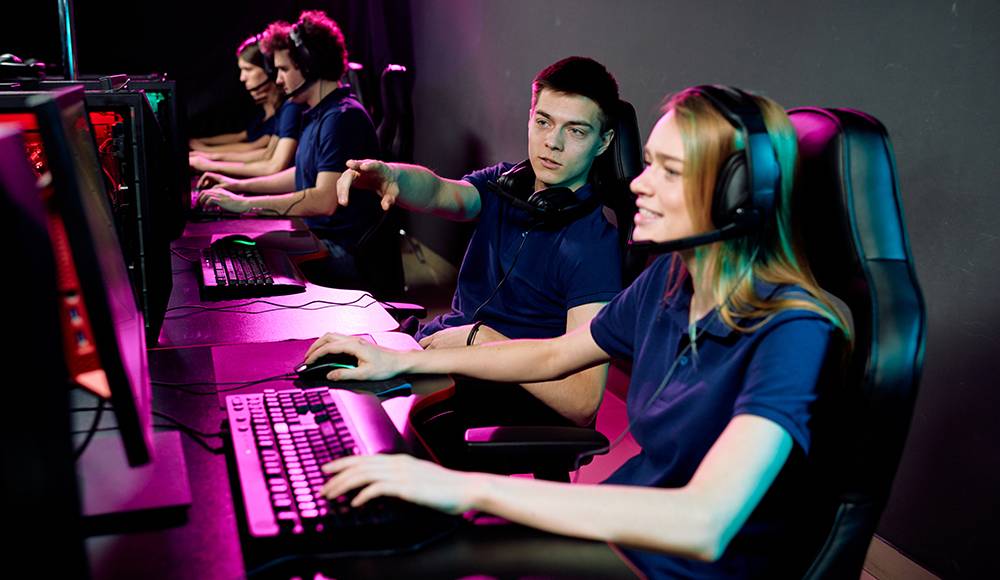 A group of gamers in a dimly lit room, focused on their screens, wearing headsets and using gaming keyboards.