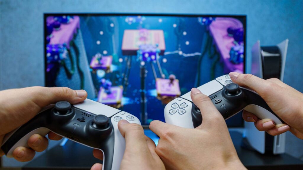 Two hands hold PlayStation 5 controllers, focused on a colorful game displayed on a TV screen in the background.