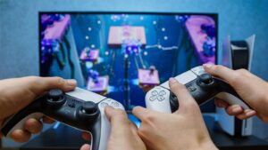 Two hands hold PlayStation 5 controllers, focused on a colorful game displayed on a TV screen in the background.