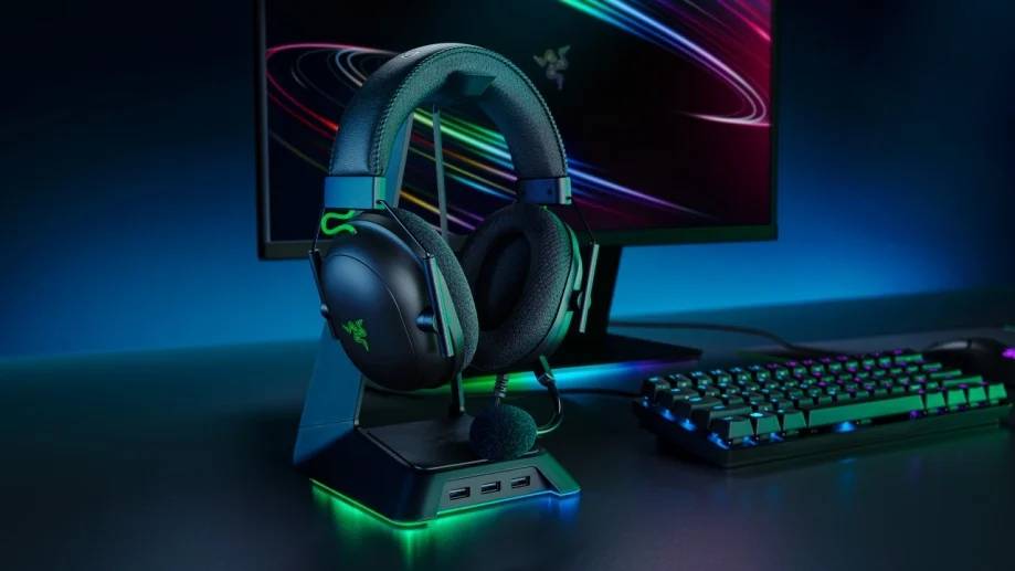  A Razer gaming headset on a stylish stand, next to a keyboard, with a vibrant monitor displaying colorful graphics in a dark setting.