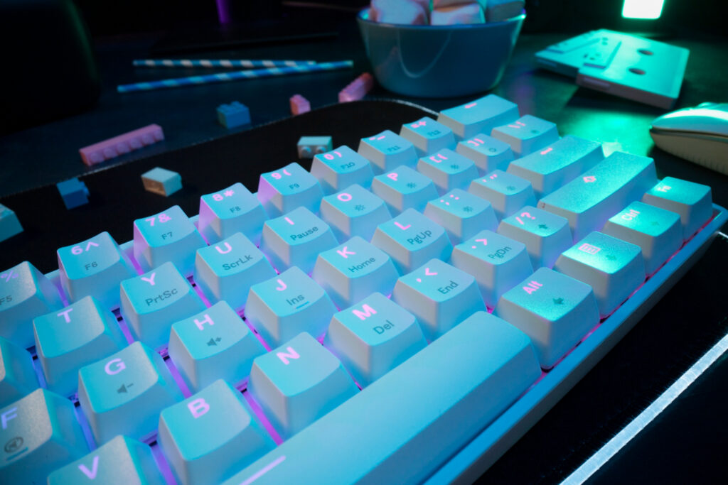 A close-up of a backlit mechanical keyboard with soft blue and purple lighting, showcasing white keys and colorful desk accessories.
