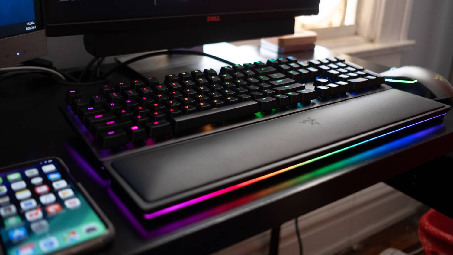 A close-up of a backlit mechanical keyboard on a desk, featuring RGB lighting, alongside a smartphone and two computer monitors.