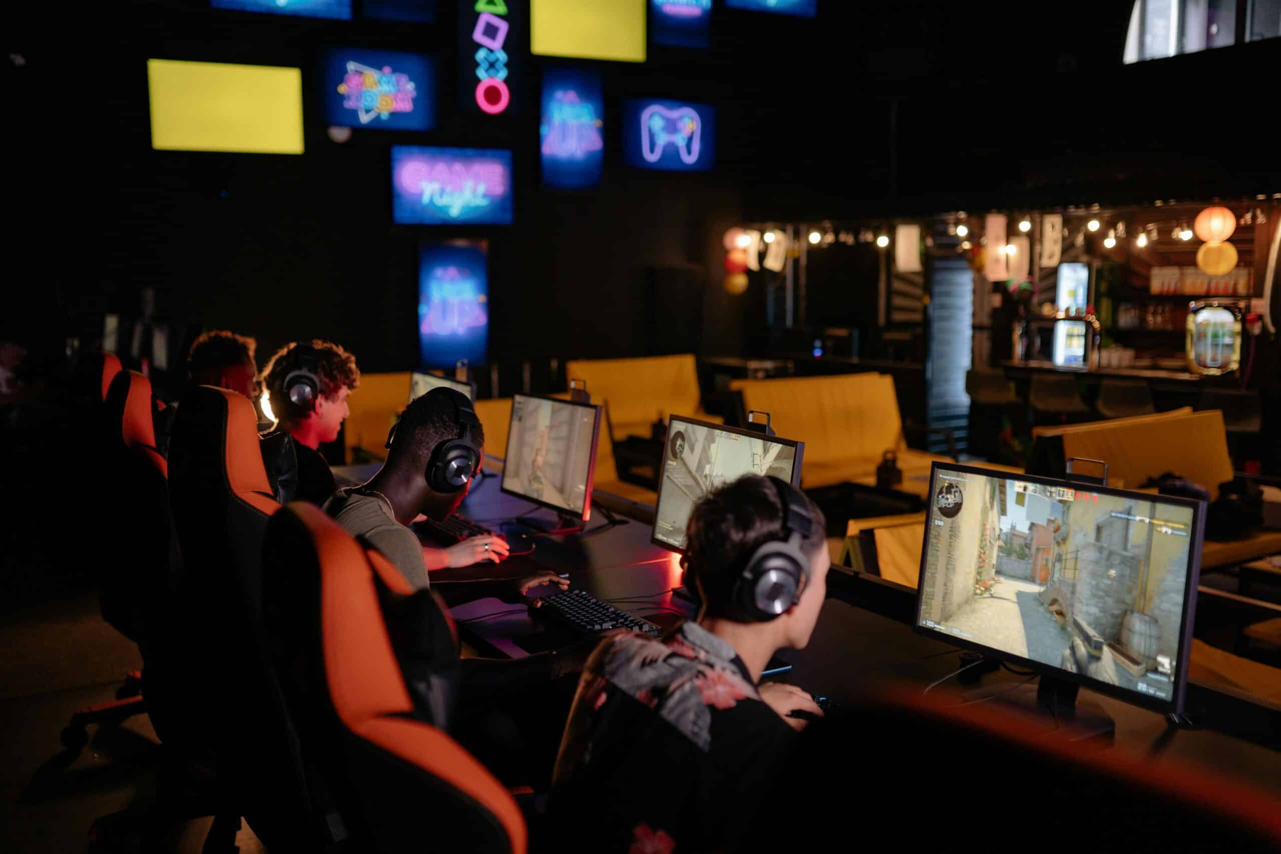 A group of gamers in a dark room, focused on their screens, surrounded by vibrant gaming-themed neon lights and cozy seating.