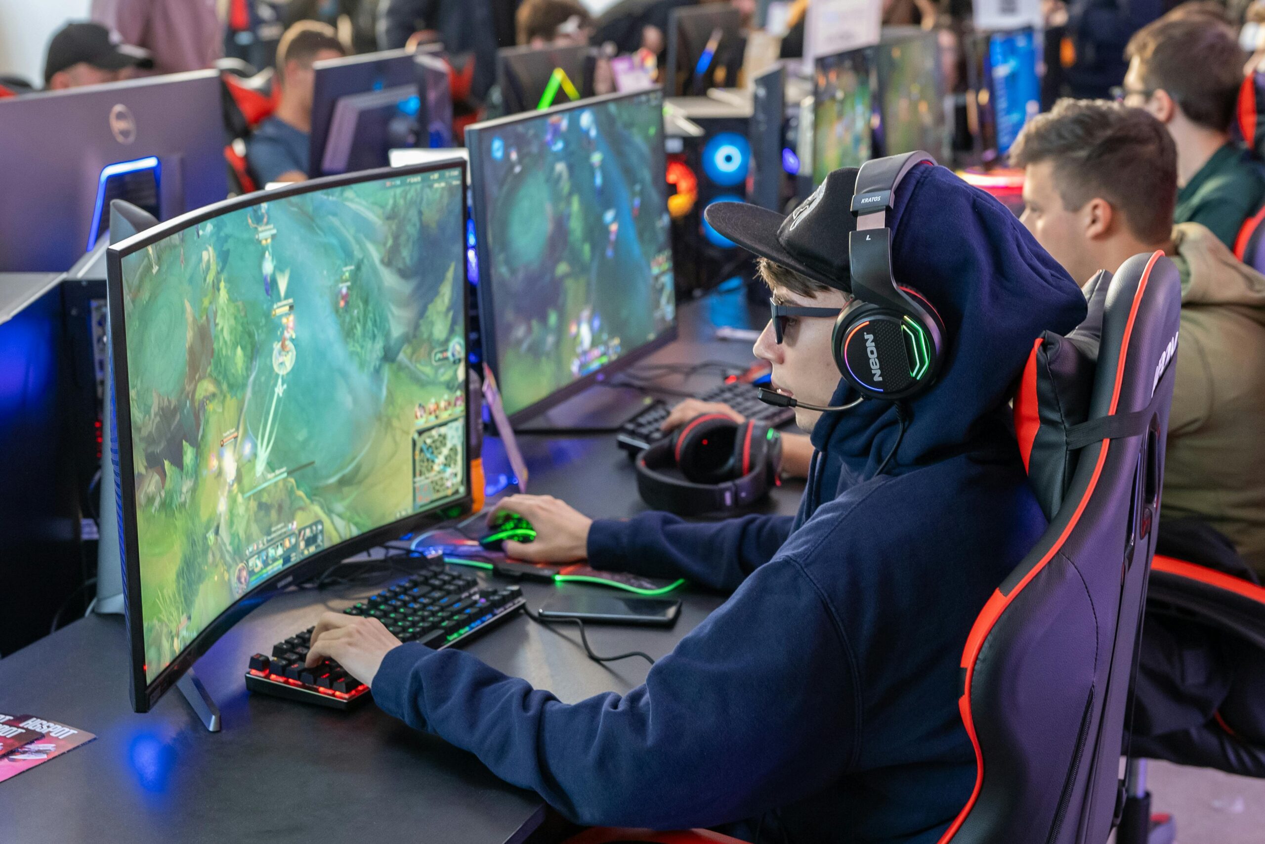  A gamer in a hoodie plays on a dual-curved monitor, surrounded by colorful gaming equipment in a lively environment.