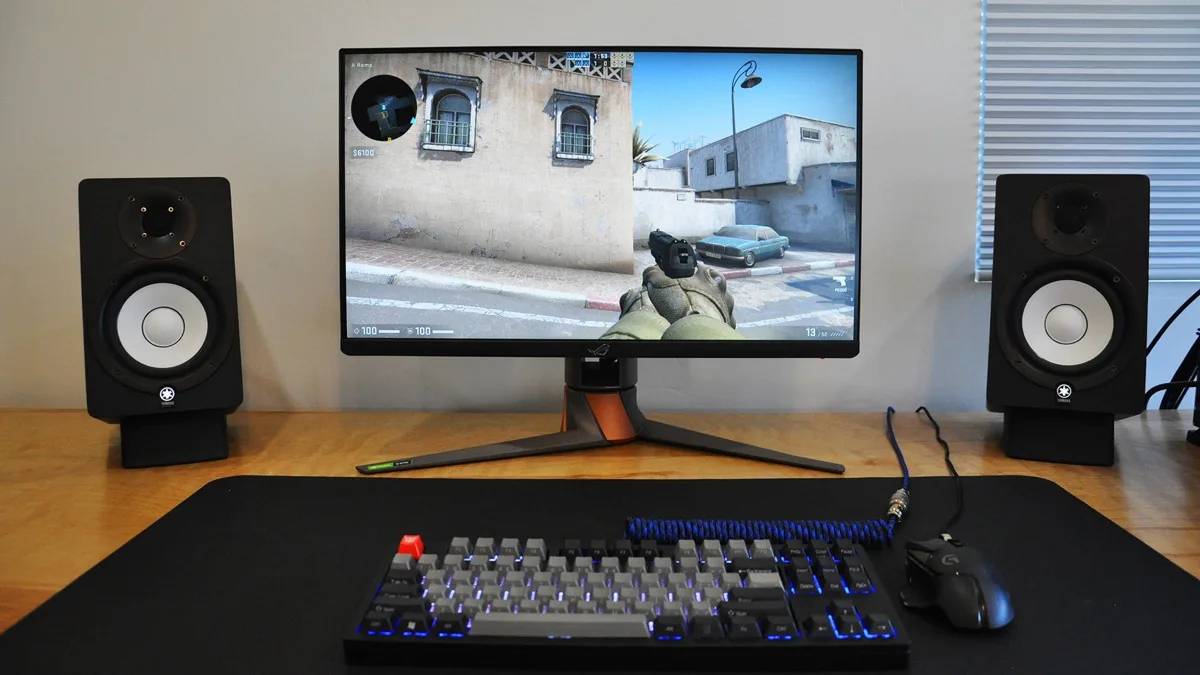  A gaming setup featuring a monitor displaying gameplay, a mechanical keyboard, a mouse, and audio speakers on a wooden desk.
