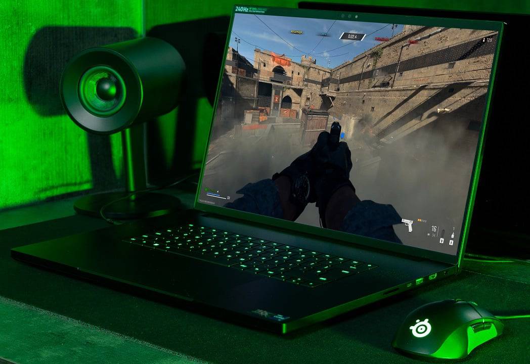 A gaming laptop displays an action scene, with a character aiming a gun, surrounded by smoky ruins; illuminated by green ambient light.