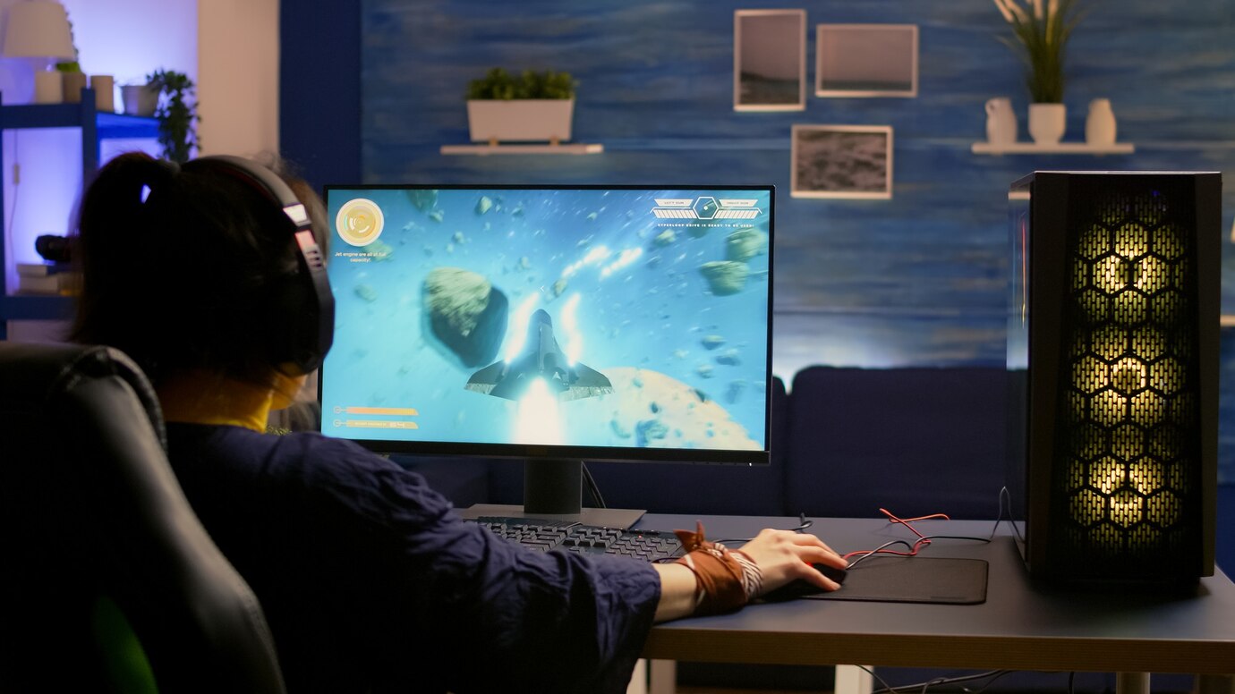 A gamer in a dark room, wearing headphones, intensely plays a space game on a monitor with colorful graphics, a gaming PC nearby.