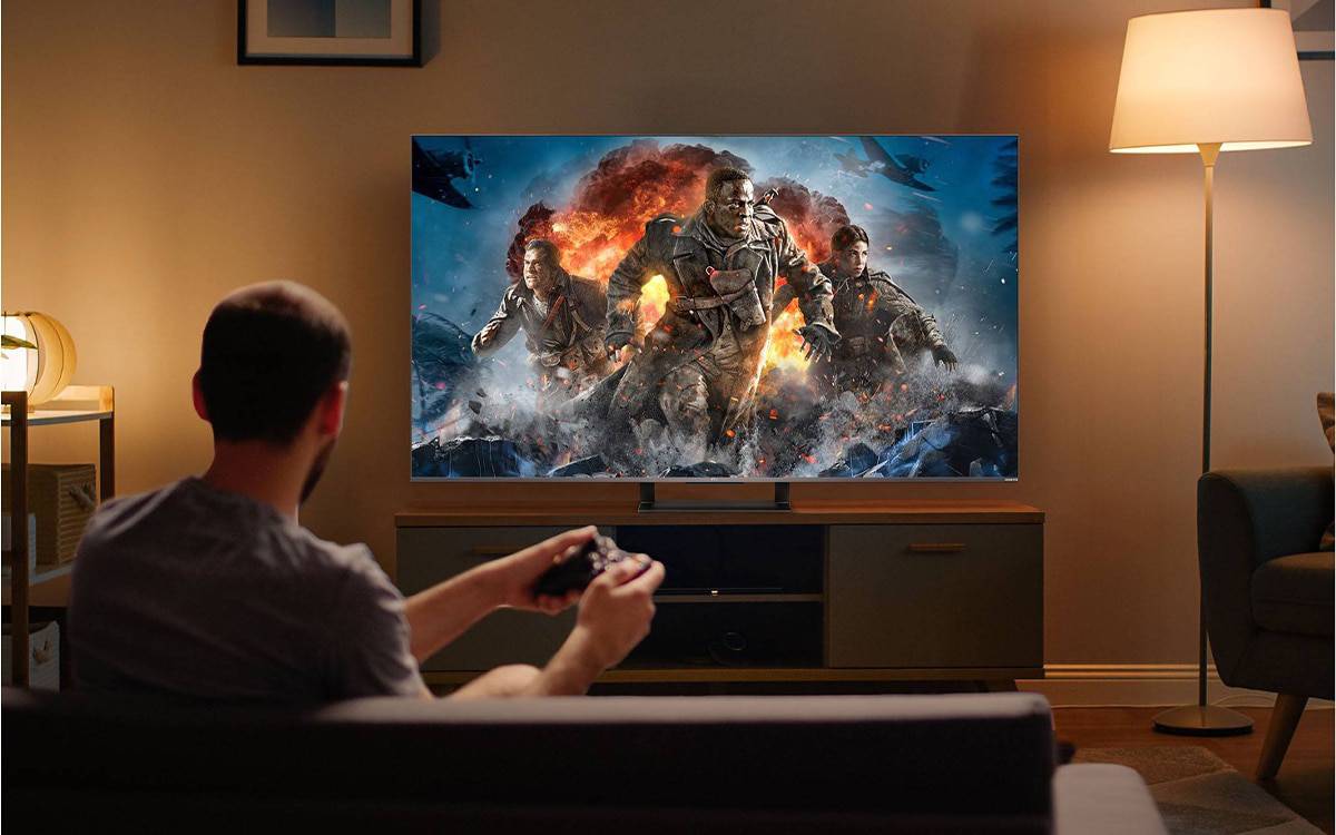 A person seated on a couch plays a video game, focused on a dramatic scene of soldiers amidst explosions displayed on a large TV.