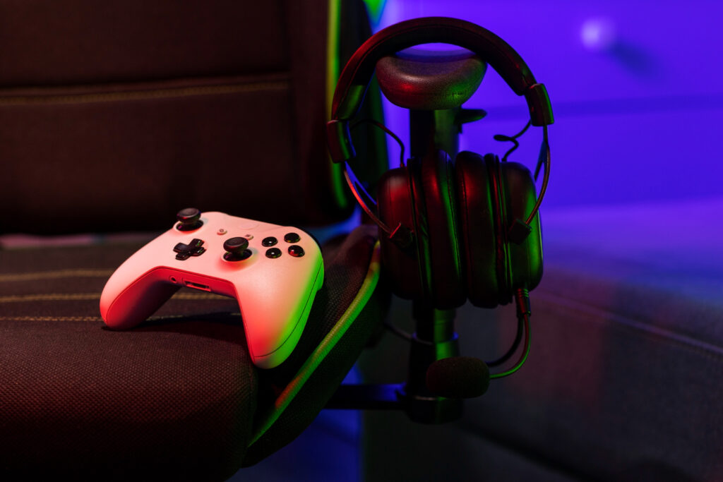 A white gaming controller rests on a gaming chair, beside a black headset with a microphone, illuminated by colorful lights.