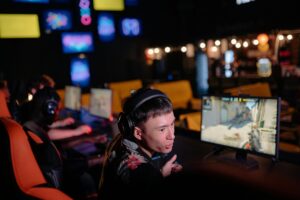 A gamer sits in an esports venue, focused on their monitor while wearing headphones, with other players visible in the background.