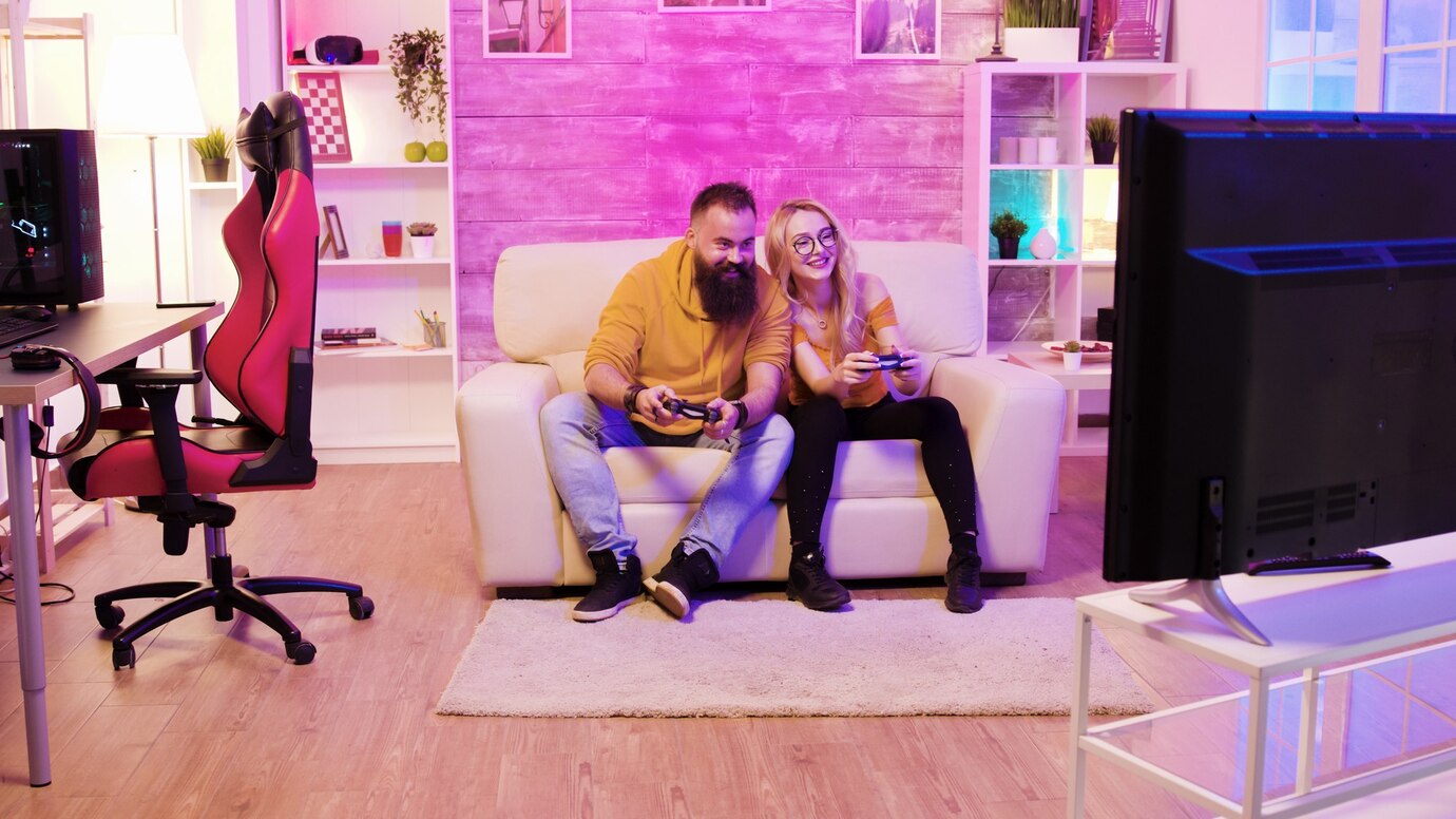 A couple sits on a white sofa, playing video games with controllers, in a cozy, colorful room with gaming equipment and vibrant decor.