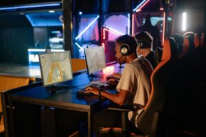 Gamers sitting at computer stations in a vibrant eSports arena, focused on their screens, wearing headphones. Colorful LED lights illuminate the space.