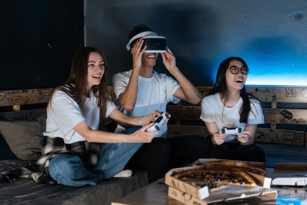 A group of three friends wearing matching white t-shirts enjoy gaming together on a couch, with pizza boxes on the table.