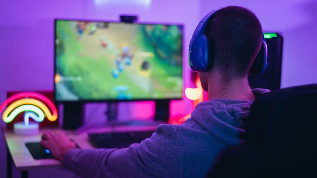A gamer wearing headphones sits in front of a colorful screen, focused on gameplay, with neon lights illuminating the space.