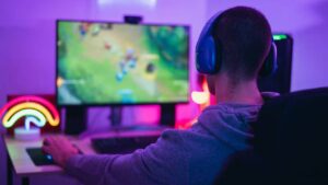 A gamer wearing headphones sits in front of a colorful screen, focused on gameplay, with neon lights illuminating the space.