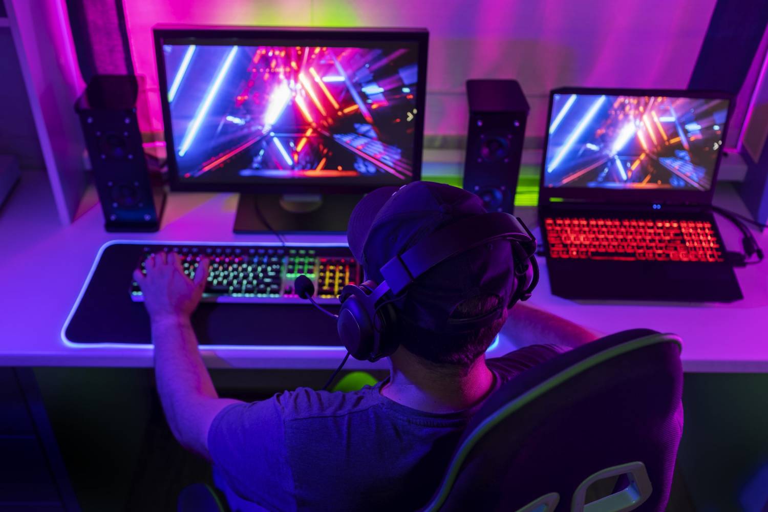 A gamer with headphones sits in a dimly lit room, focused on dual screens displaying colorful visuals and gaming graphics.