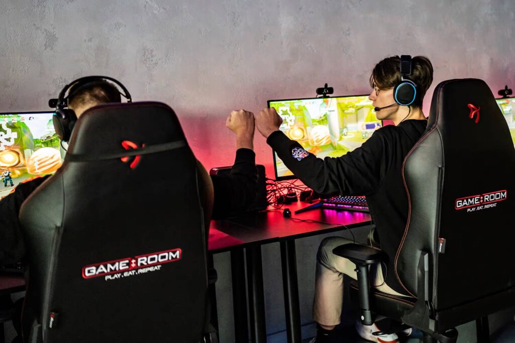 Two gamers celebrate a victory with raised fists, seated at gaming desks with colorful monitors and high-end gaming chairs.
