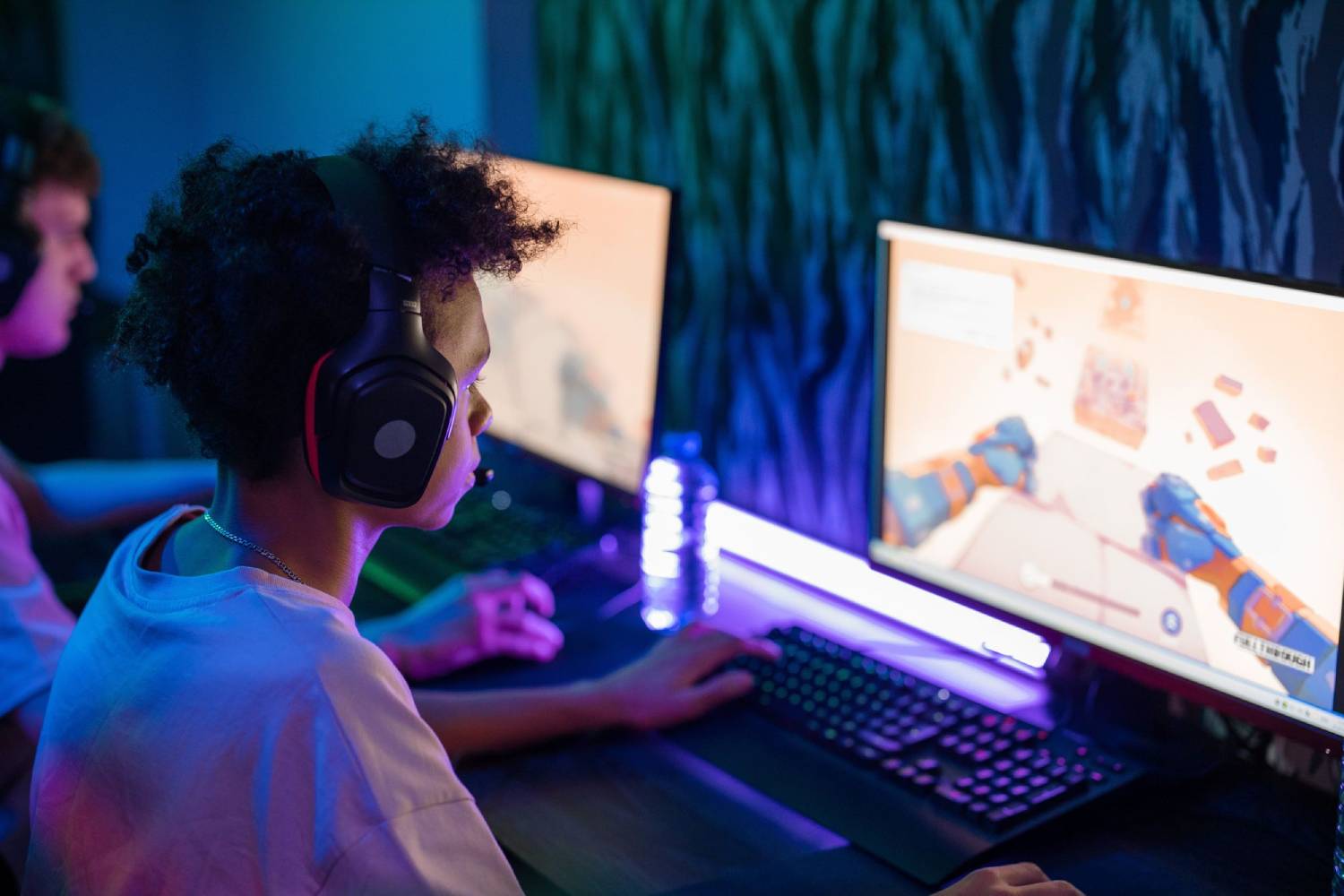 A gamer wearing headphones plays on a computer, focused on a colorful game displayed on the monitor, with a glowing water bottle nearby.