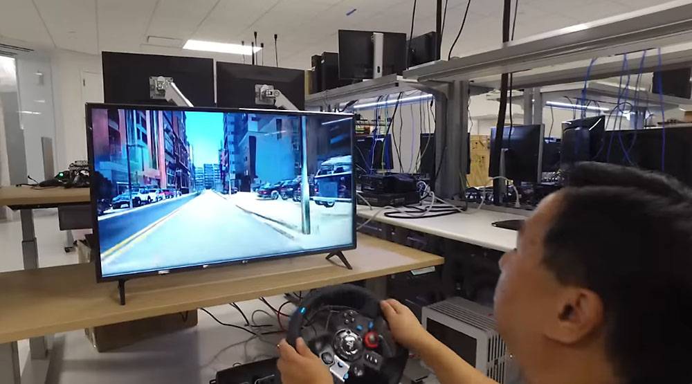 A person drives a racing simulator with a steering wheel, while a large screen displays a city street scene ahead.