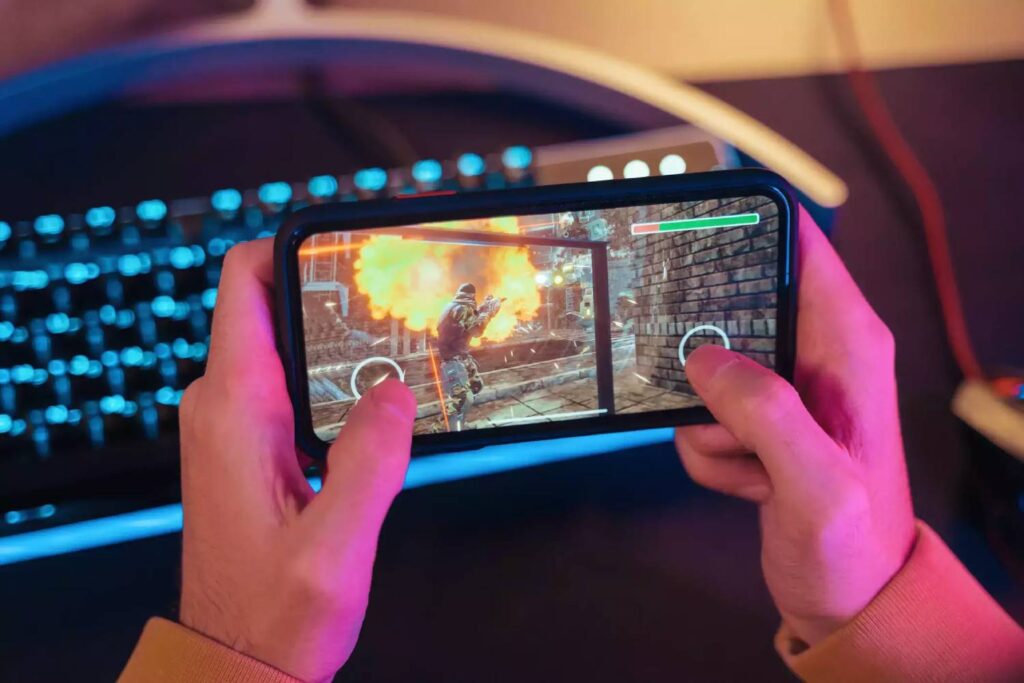 A person holds a smartphone, playing a shooting game with a fiery explosion on the screen, surrounded by a glowing keyboard.