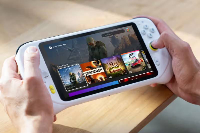 A person holds a handheld gaming device displaying a game library, featuring titles like Deathloop and Forza.