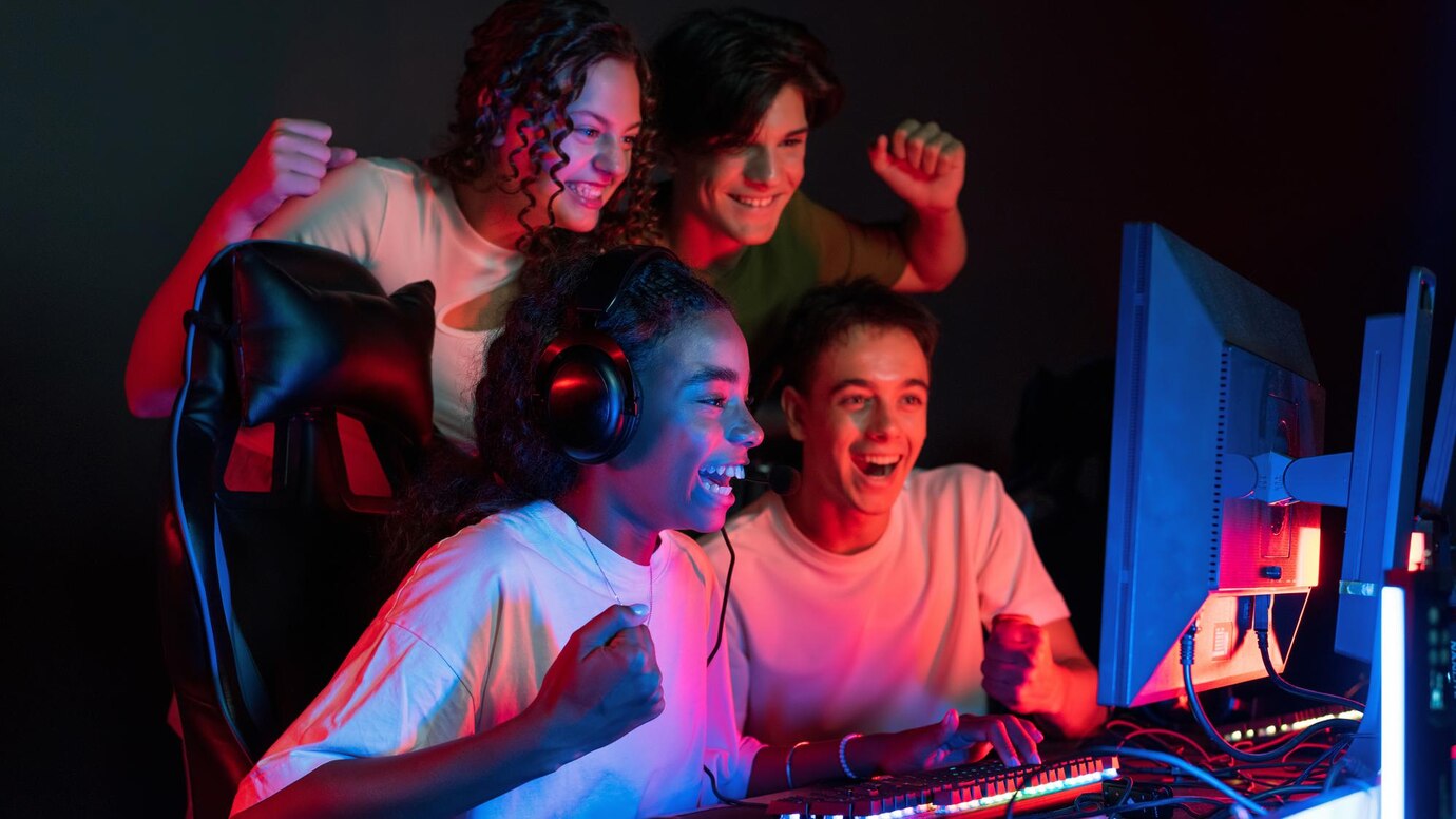 A group of enthusiastic gamers celebrates a win, sitting at a computer desk illuminated by vibrant red and blue lights.