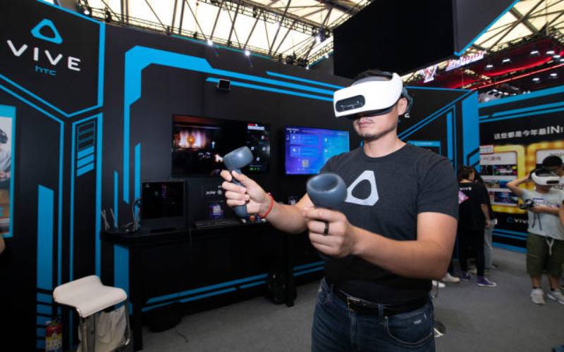 A person engages with HTC Vive virtual reality equipment at a tech event, surrounded by displays and fellow attendees.