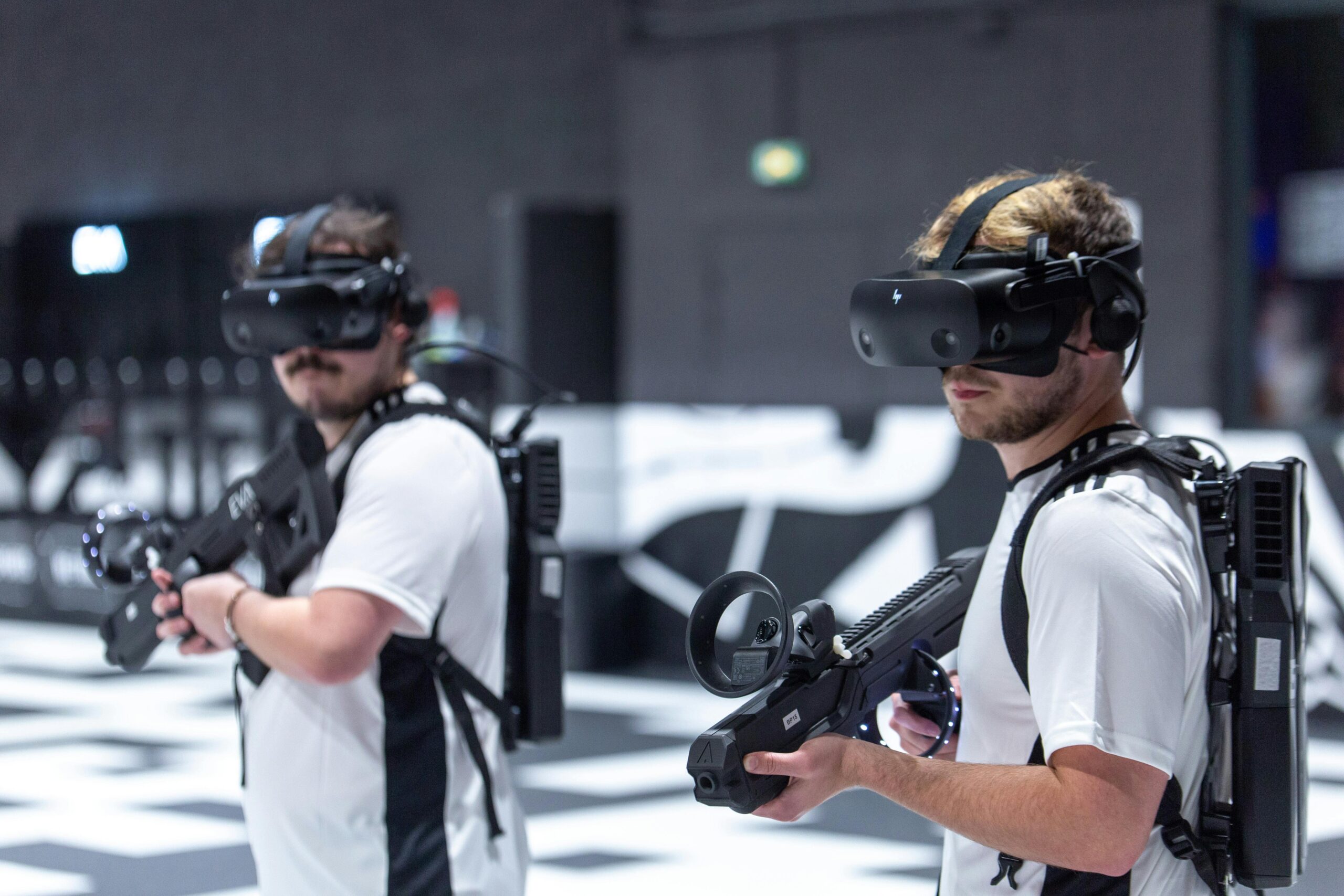 Two individuals wear VR headsets and tactical gear, holding futuristic weapons in a high-tech gaming arena.