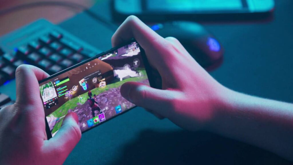 A person holding a smartphone displays a Fortnite gaming screen, set against a backdrop of a keyboard and mouse in dim light.