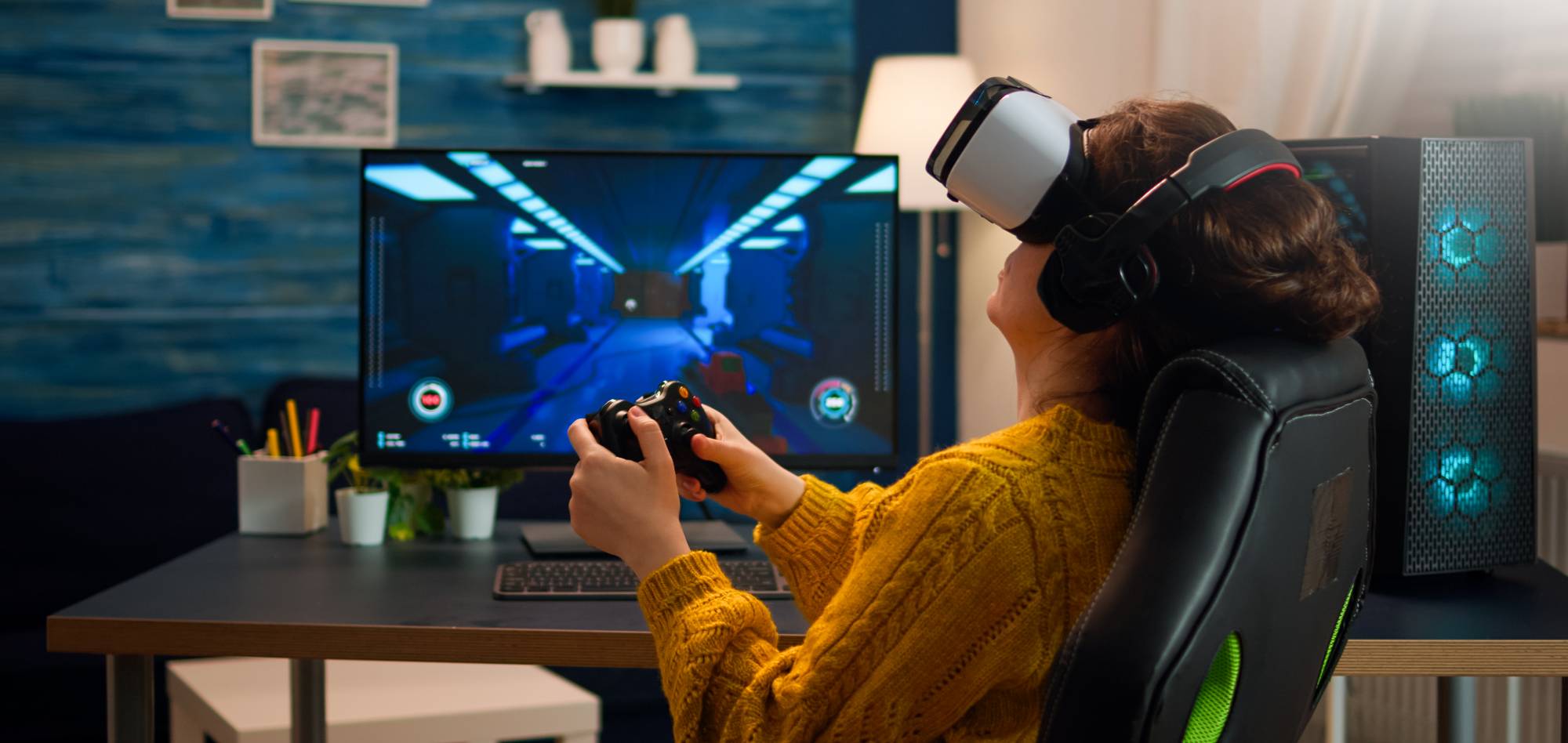 A person in a yellow sweater wears a VR headset, immersed in gaming, with a controller in hand and a computer screen displaying a game.