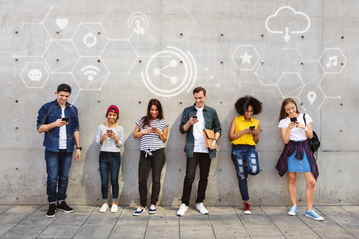 Group of young adults using smartphones against a wall with digital icons and symbols floating above them.