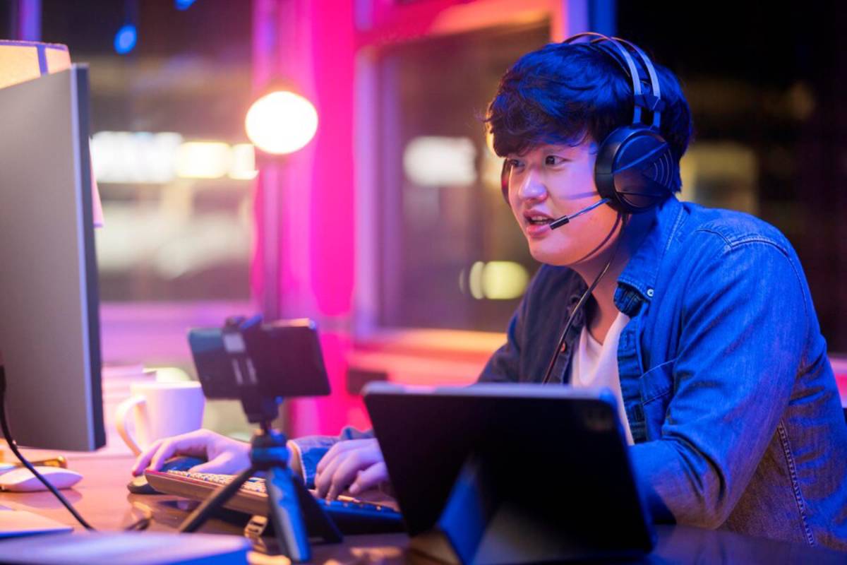 Young Asian man with headphones using multiple electronic devices at a neon-lit desk.