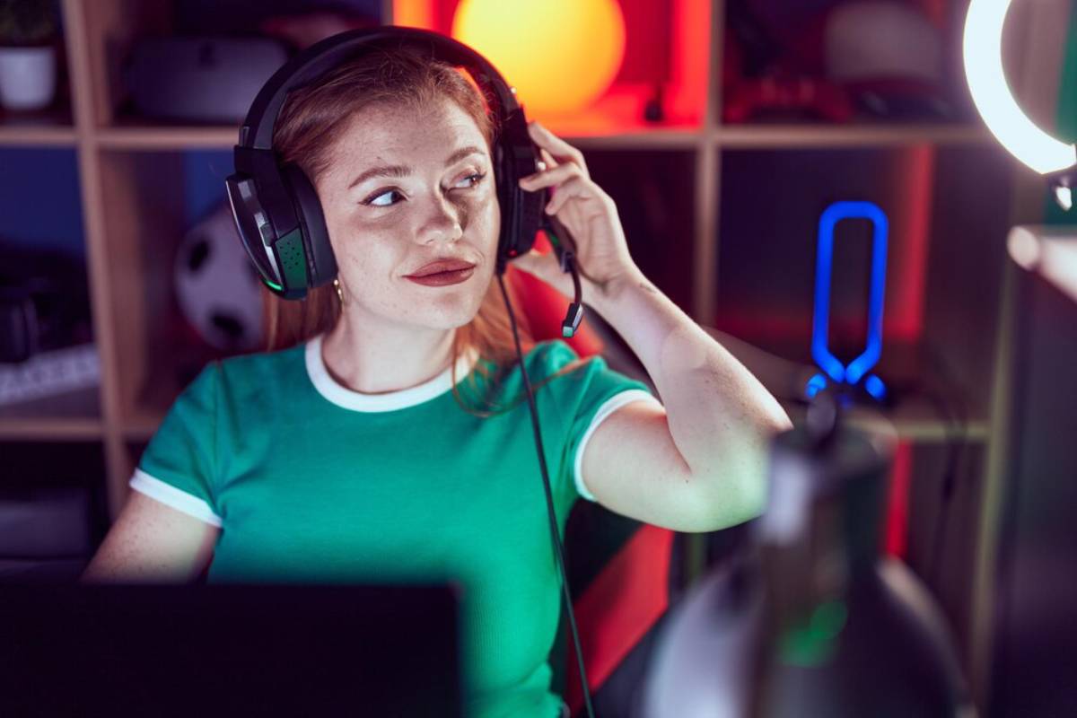 Young woman in a gaming setup wearing headphones and adjusting microphone, with colorful lighting in the background.