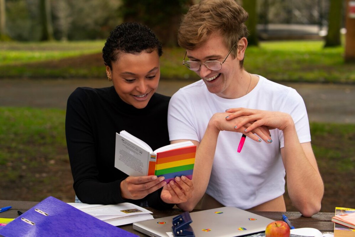 Two students reading and smiling at a rainbow-colored book outdoors at a park table, surrounded by study materials.