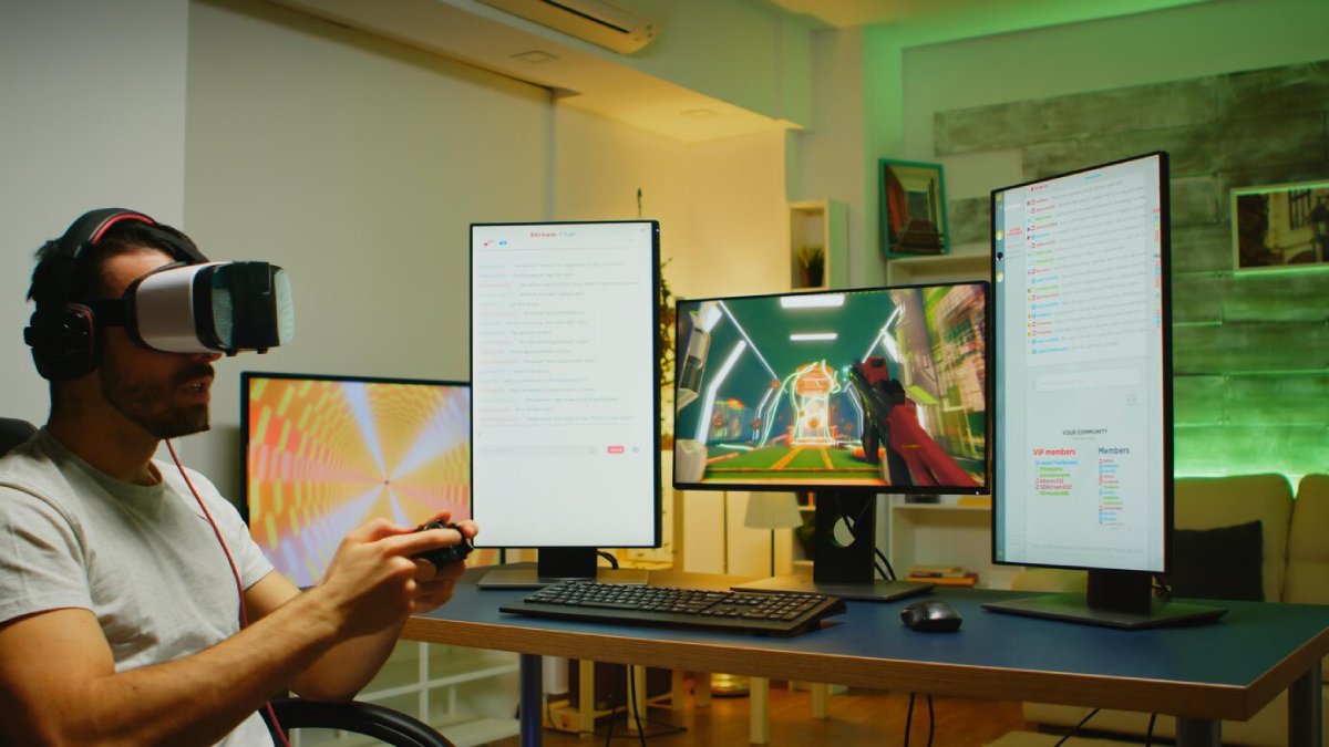 Man using virtual reality headset and controller in a home office with multiple computer screens displaying graphics and data.