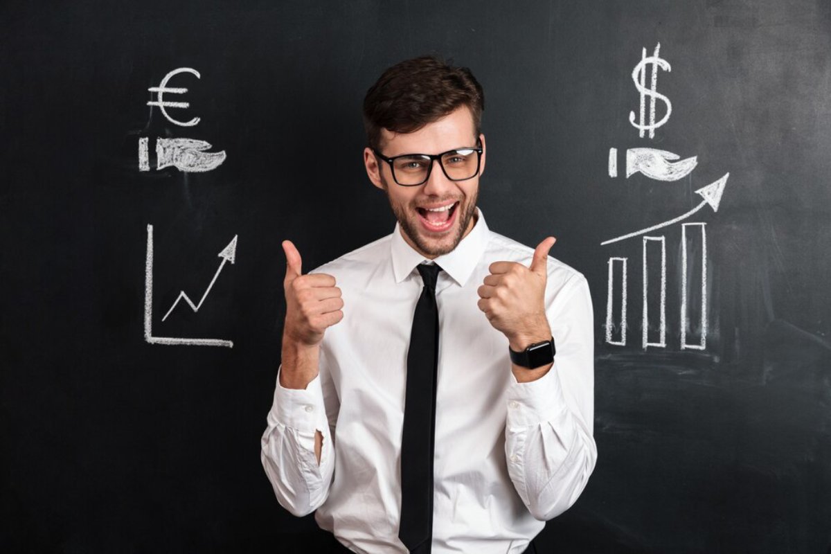 A cheerful businessman giving thumbs up in front of a chalkboard with financial symbols and graphs.