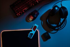 Gaming setup with illuminated keyboard, mouse, headset, and gadgets on a desk with blue lighting.