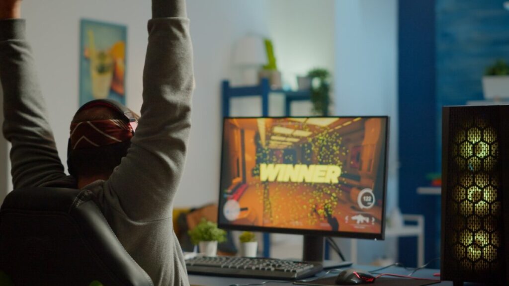 Person in headphones celebrating a win on a computer game screen in a cozy, well-lit room.
