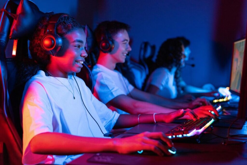 Three young gamers wearing headsets at illuminated computer stations in a dark room, focused on screens.