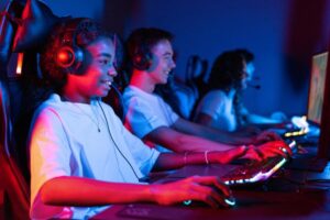 Three young gamers wearing headsets at illuminated computer stations in a dark room, focused on screens.